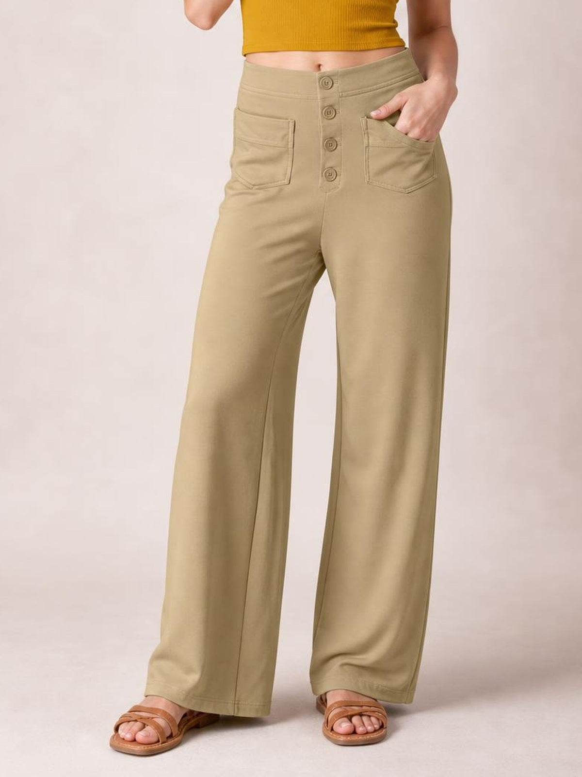 A model stands in taupe high-waist wide-leg pants with a front button fly, belt loops, stitched patch pockets, and one hand tucked into a pocket.