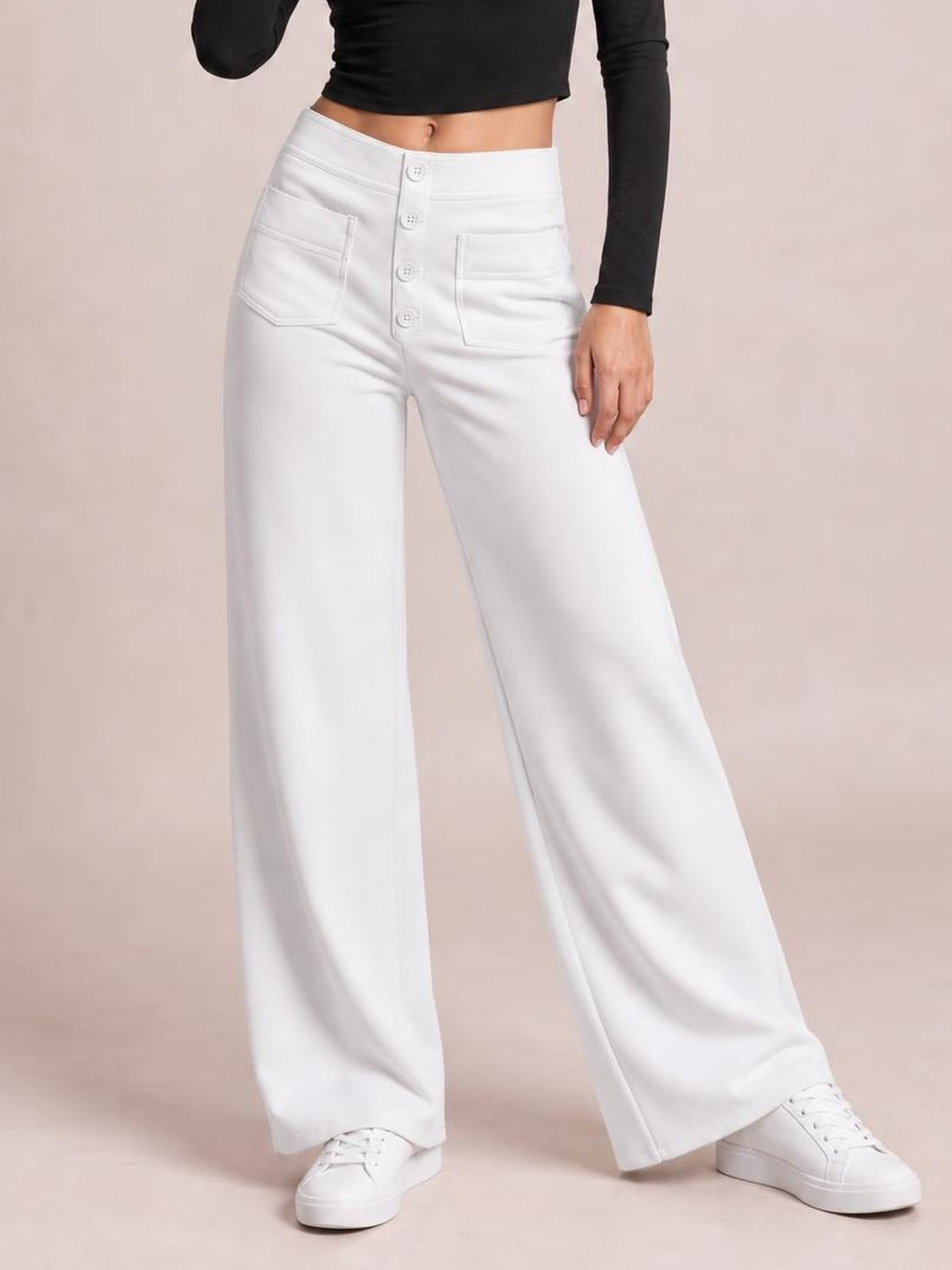 A model poses in opaque white high-waist wide-leg pants with front pockets and a button-front closure, styled with a black cropped top and sneakers.