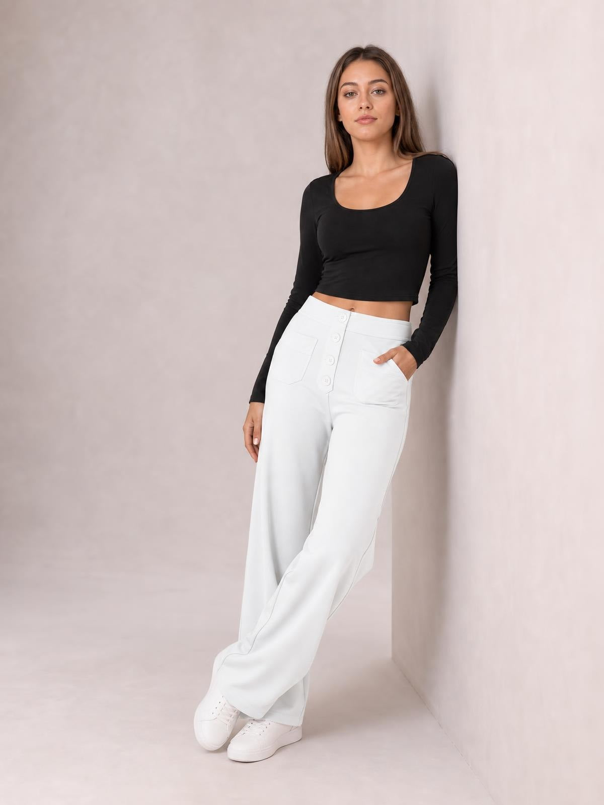 Model poses in white high-waist wide-leg pants with front pockets and button-fly construction, one hand tucked into a pocket and legs crossed.