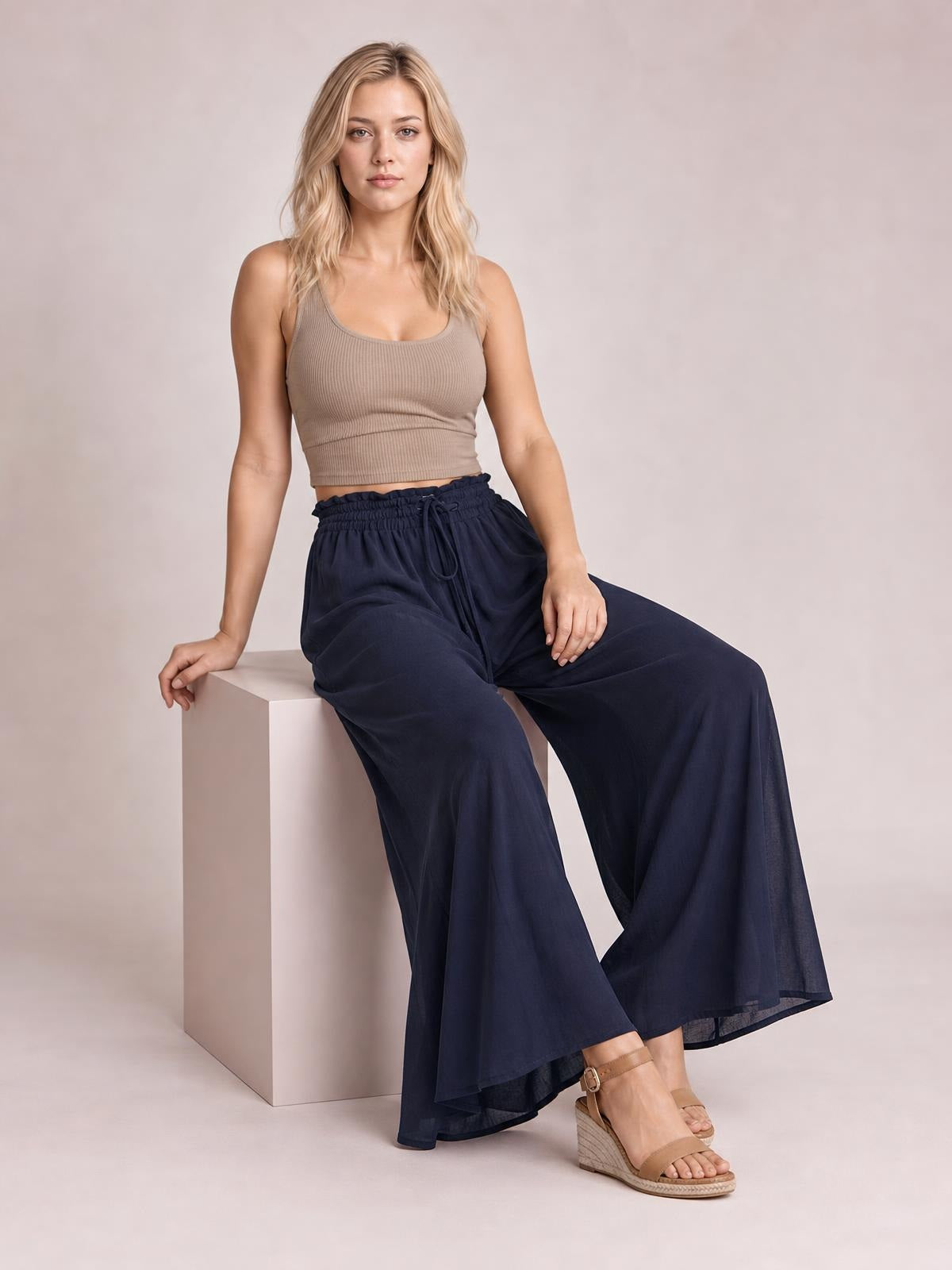A seated model wears navy high waist wide leg pants with a relaxed flowing silhouette, gathered drawstring waistband, and side pockets.
