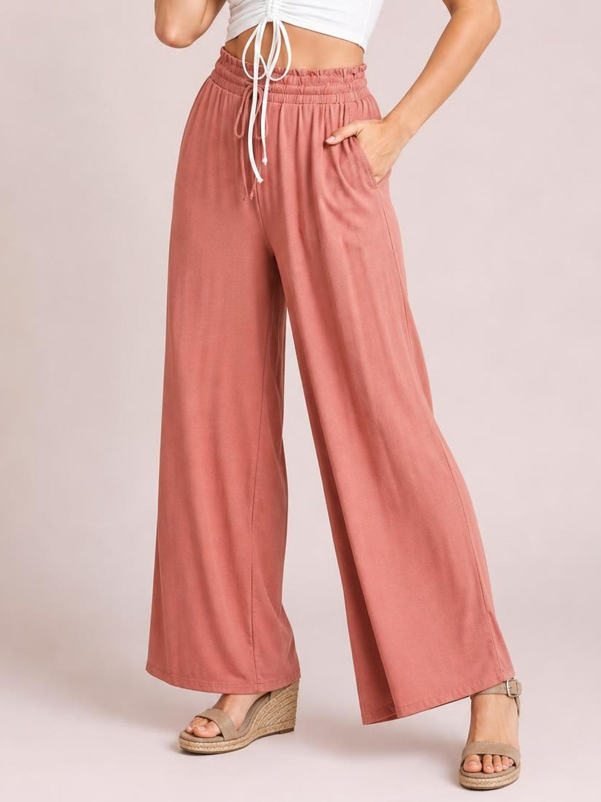 Model wearing dusty rose high waist wide leg pants with an elasticized waistband, straight flowing silhouette, and side pockets, posing with one hand tucked into a pocket.