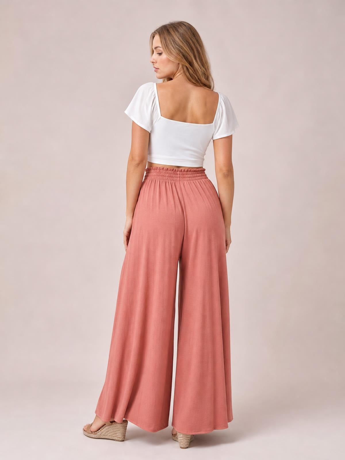 A model turns to show dusty rose high-waist wide-leg pants with a gathered elastic waistband and flowing full-length silhouette.
