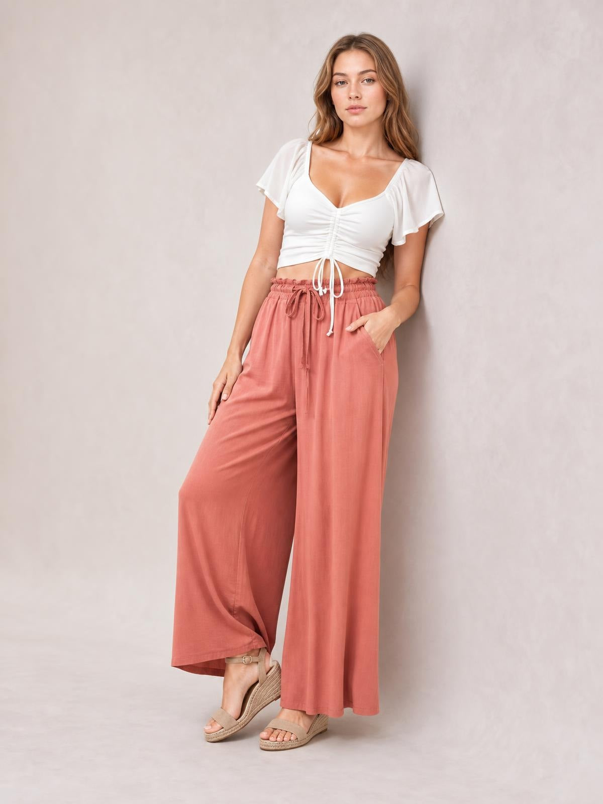 Model poses in coral high-waist wide-leg pants with an elastic drawstring waist and side pockets, one hand tucked into a pocket.