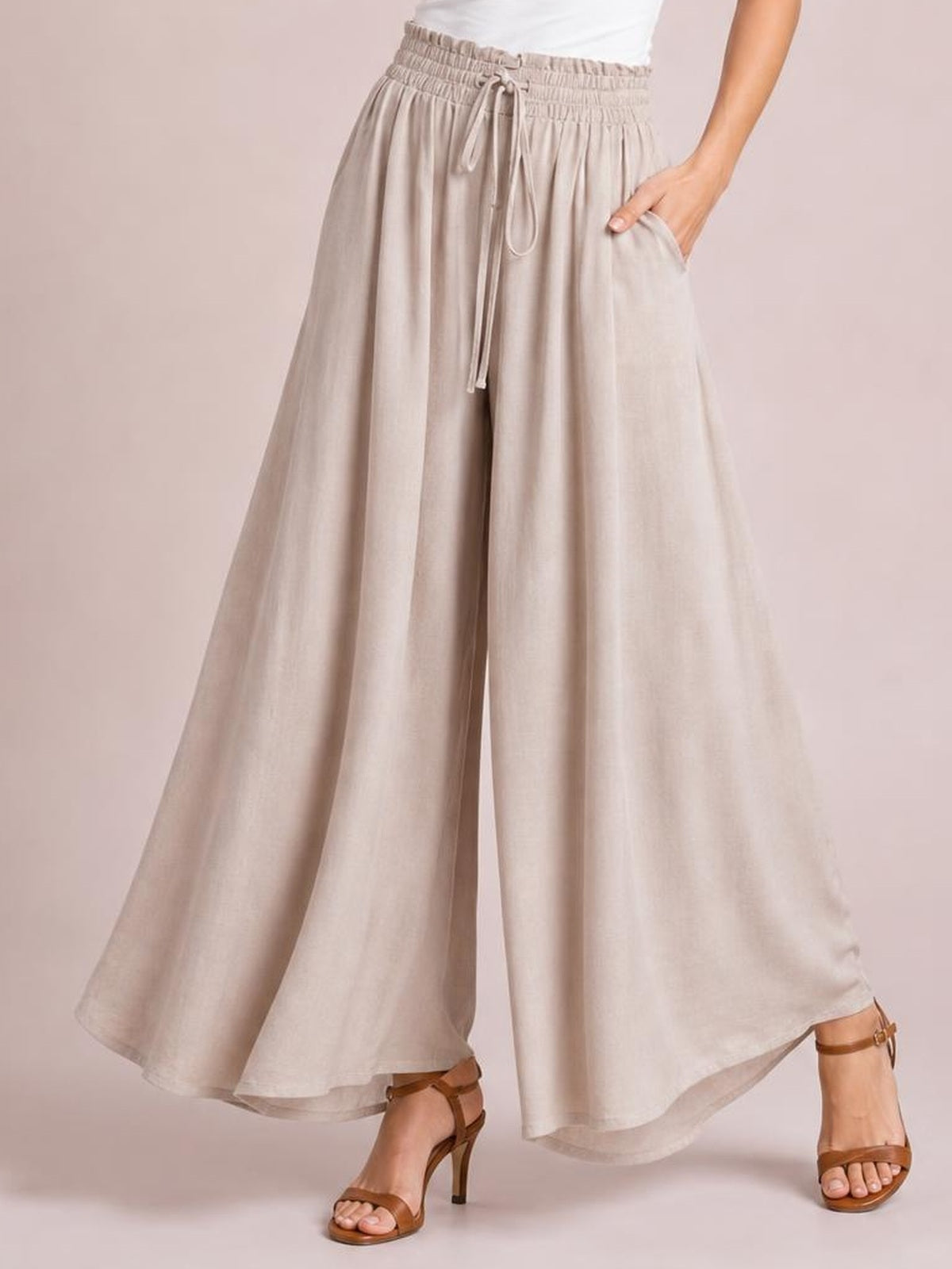 Model wearing beige high waist wide leg pants with a drawstring waistband, flowing wide-leg silhouette, and side pockets, posing with one hand tucked into a pocket.
