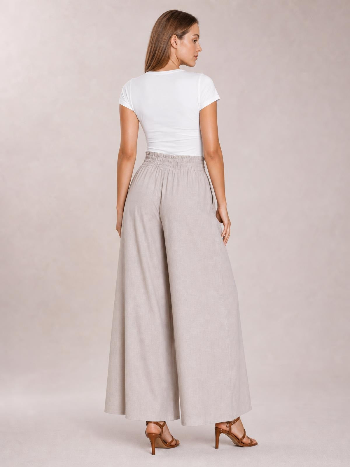 Model turns to show light beige high waist wide leg pants with an elastic waistband, flowing full-length silhouette, and side pockets.