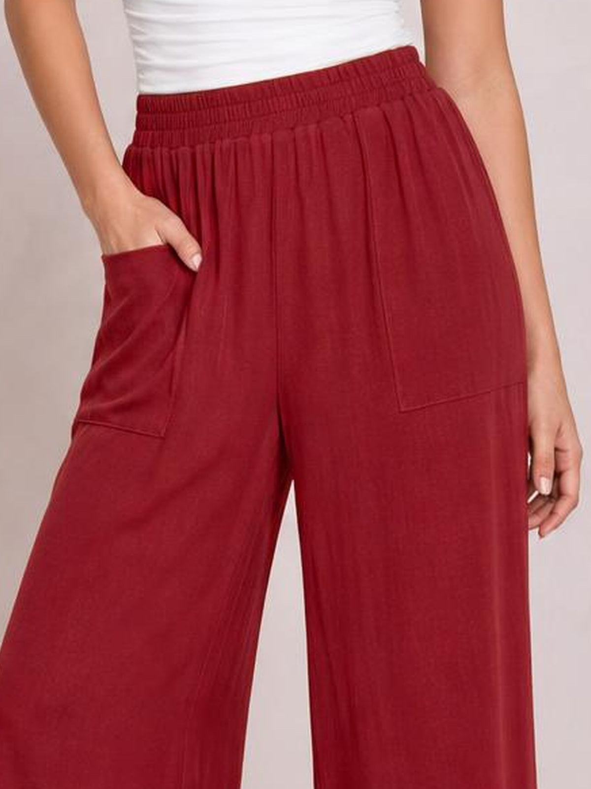 Model wearing burgundy high-waist wide-leg pants with an elastic waistband, roomy silhouette, and large stitched front pockets, one hand tucked into a pocket.