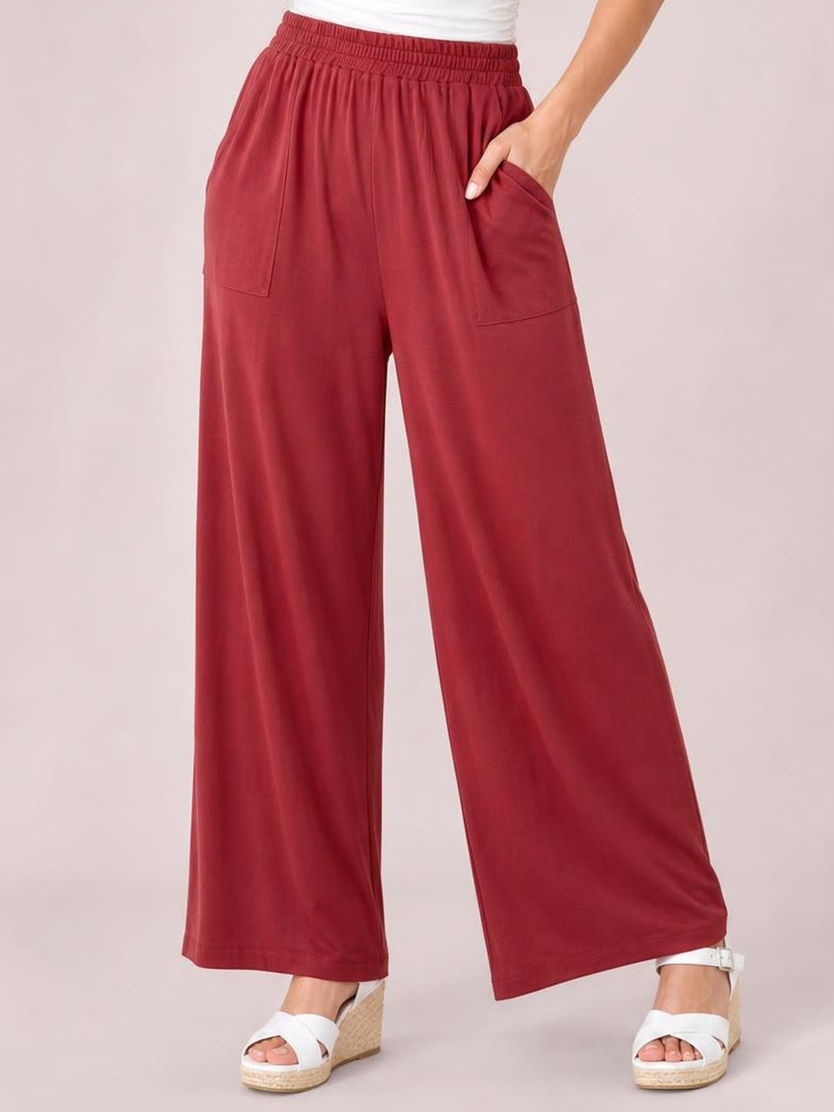 A model poses in deep red high-waist wide-leg pants with a gathered elastic waistband and visible pockets, placing one hand in a pocket.