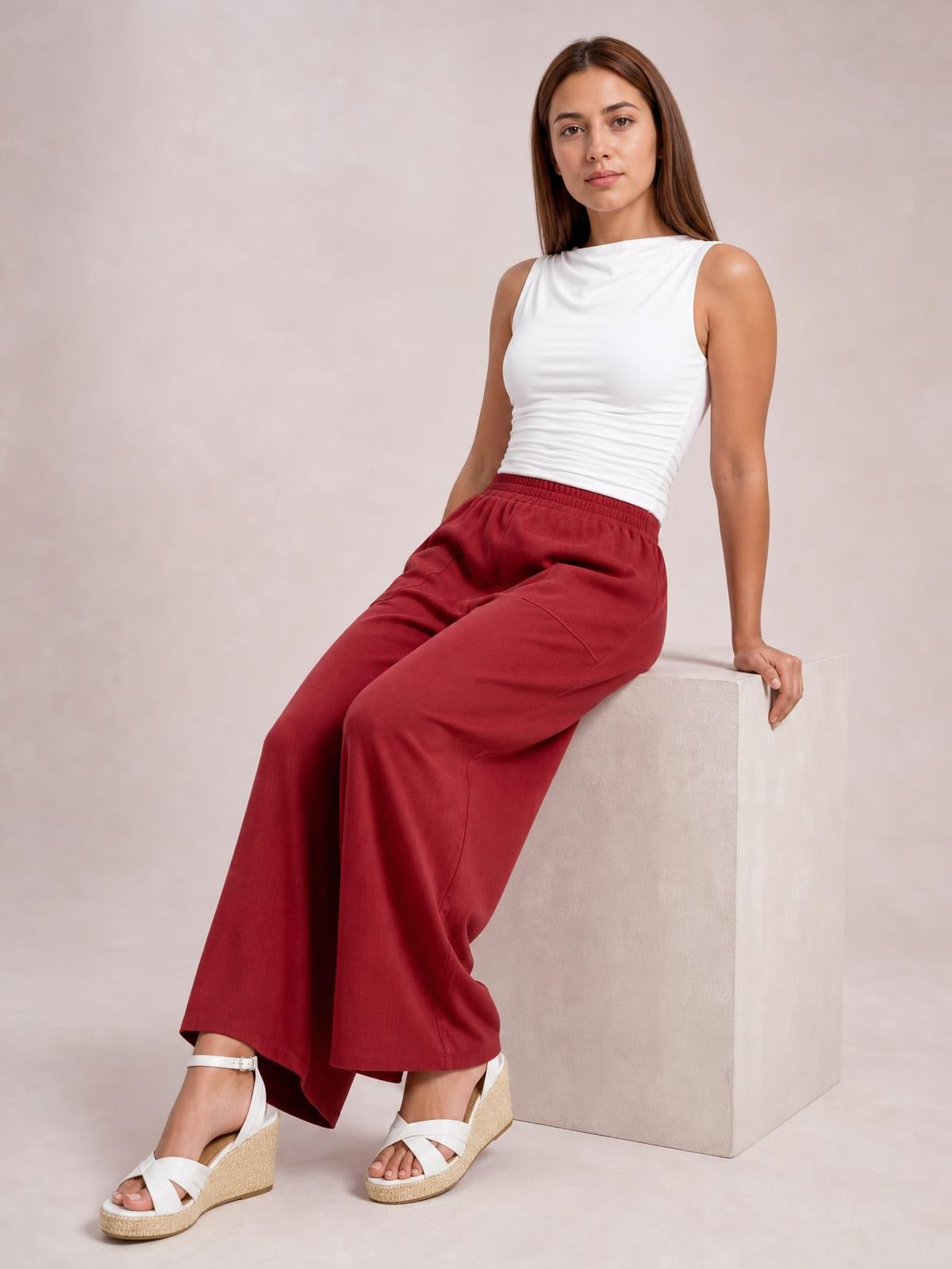A seated model wears burgundy high waist wide leg pants with an elastic waistband, roomy silhouette, and visible side pockets.