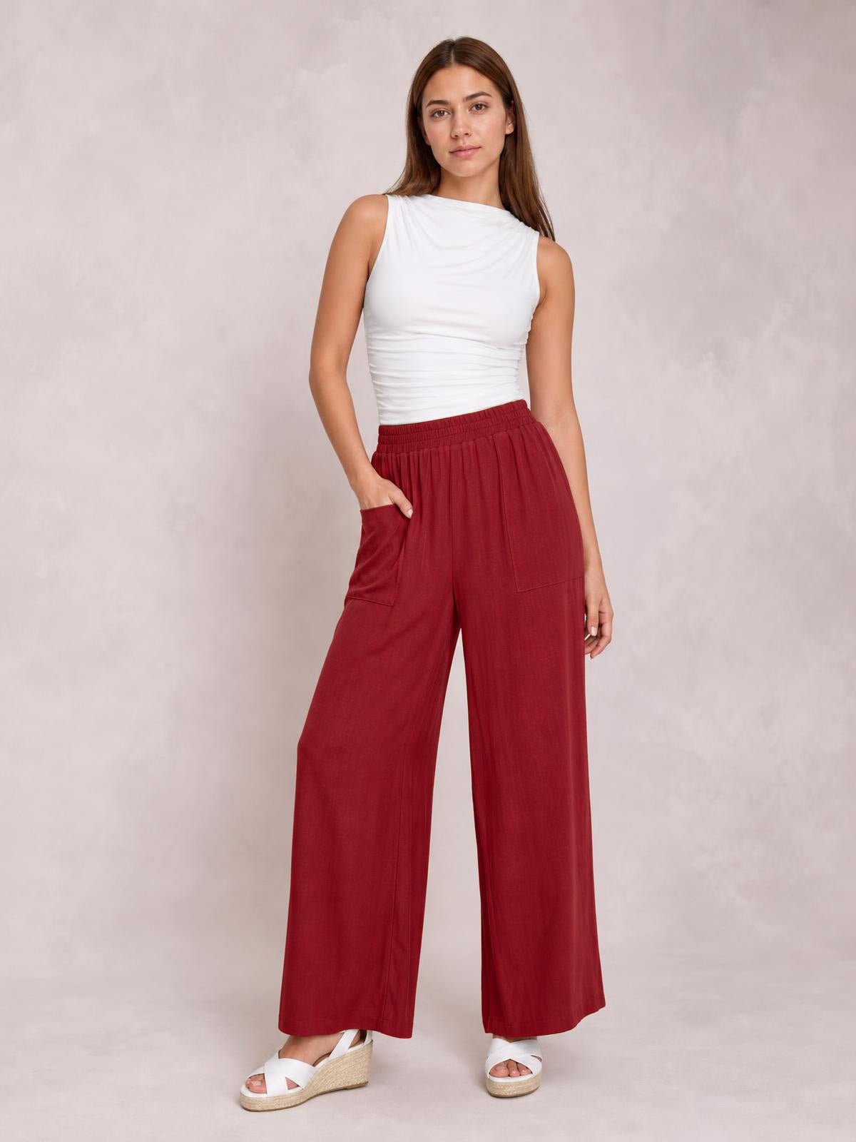 A model poses with one hand in the pocket of opaque red high-waist wide-leg pants, featuring an elastic waistband and relaxed floor-length silhouette.