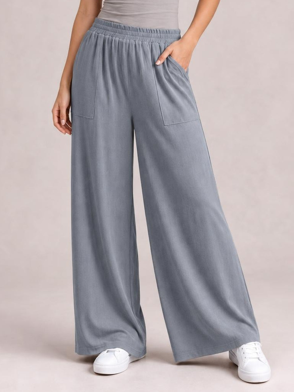 Model wearing gray high waist wide leg pants with an elastic waistband, roomy silhouette, and large front pockets, posed with one hand tucked inside a pocket.
