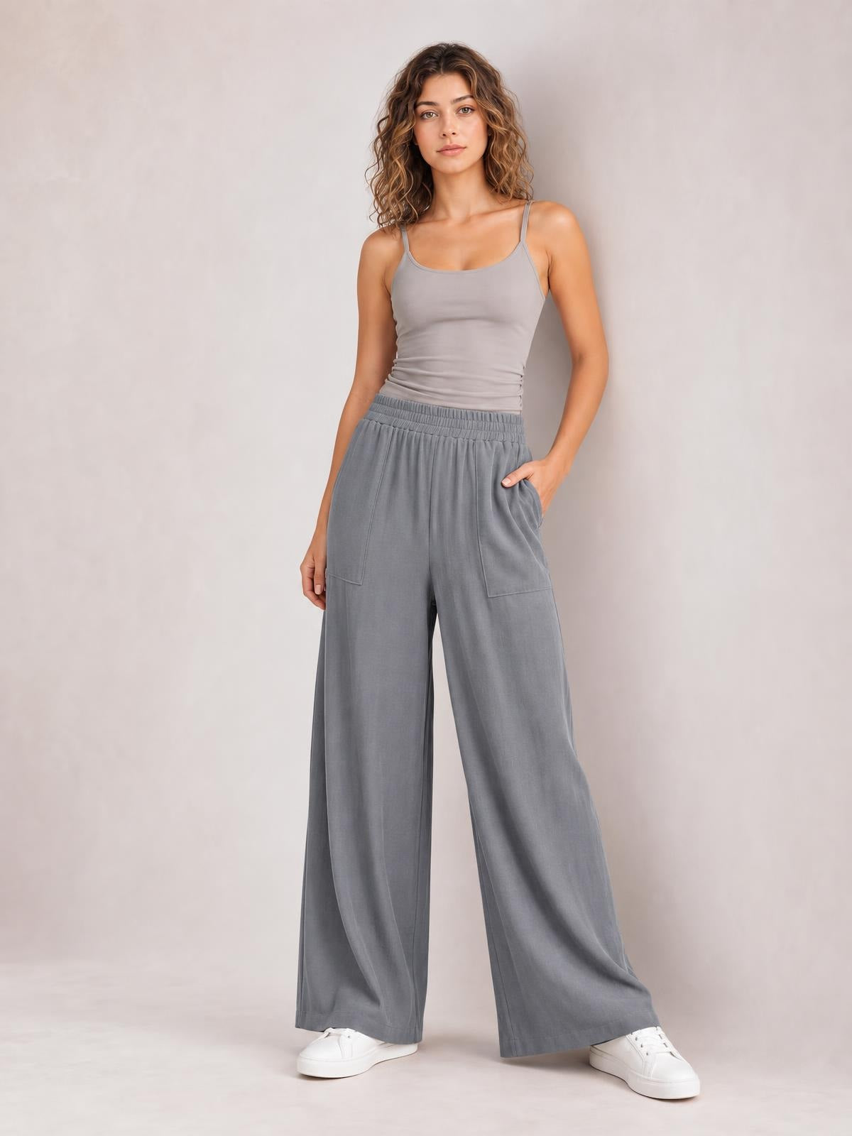 Model wears gray high-waist wide-leg pants with an elastic waistband and large front pockets, standing with one hand tucked into a pocket.