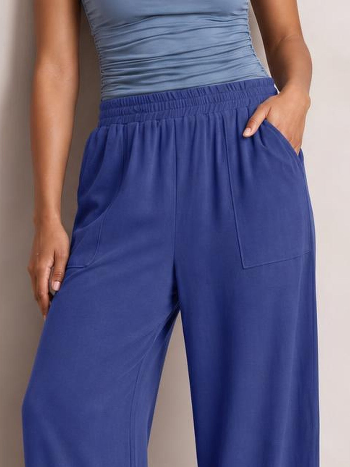 Model wearing royal blue high-waist wide-leg pants with an elastic waistband and large pockets, shown with one hand tucked into a pocket.