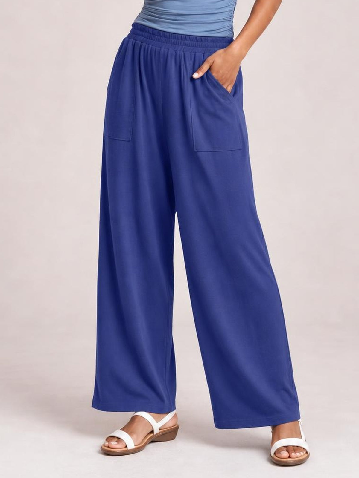 Model wearing royal blue high-waist wide-leg pants with an elastic waistband and patch pockets, standing in white sandals with one hand in a pocket.