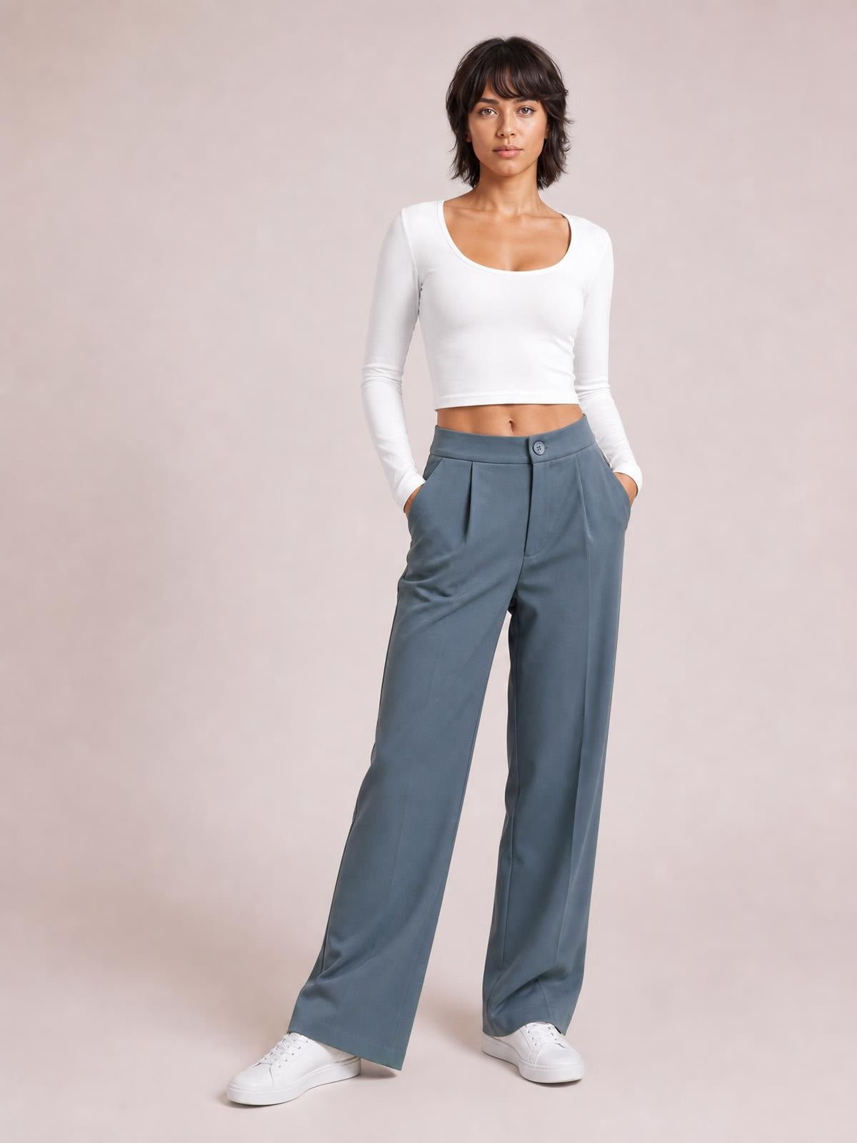 Model poses with hands in pockets wearing blue-gray high waist wide leg pleated pants with a button closure, tailored waistband, and relaxed full-length silhouette.
