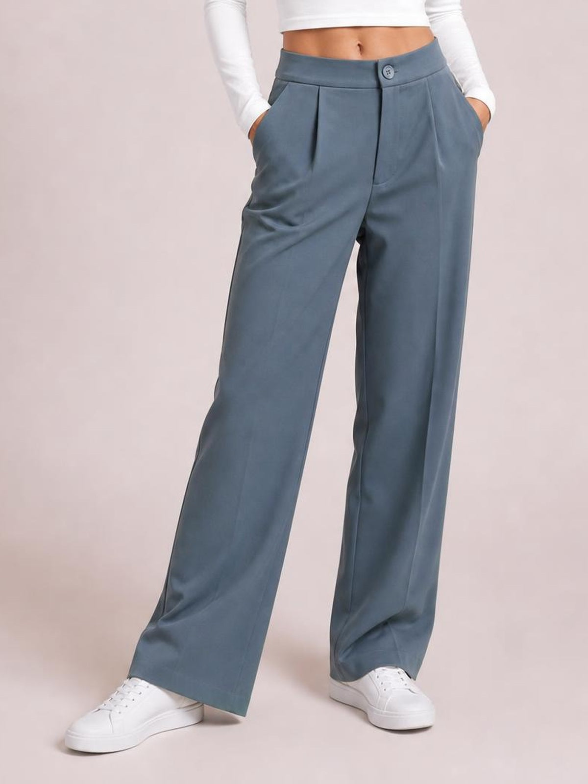 Model wearing blue-gray high waist wide leg pleated pants with a button closure and side pockets, standing with hands in pockets.