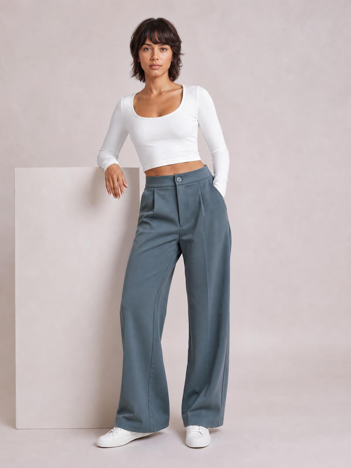 A model wears blue-gray high-waist wide-leg pleated pants with a button closure and side pockets, posed with one hand in a pocket.