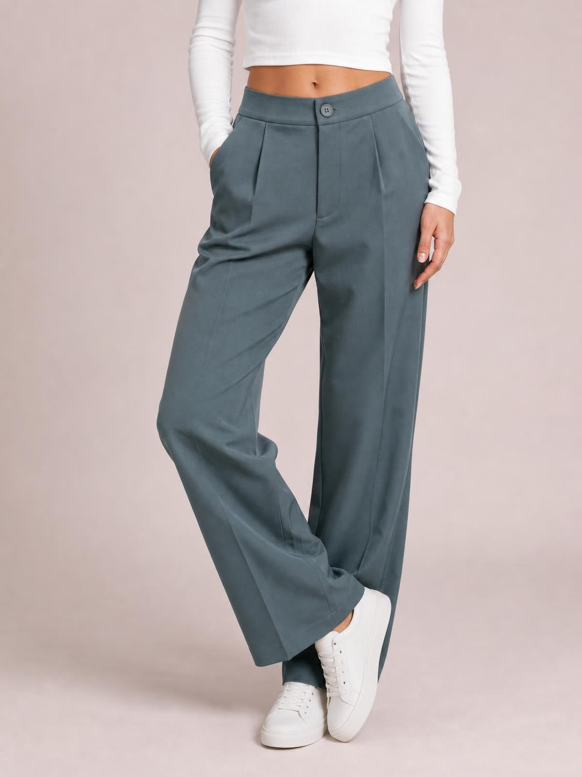 A model poses with crossed legs in slate-blue high-waist wide-leg pleated pants featuring a buttoned waistband, front pleats, and side pockets.
