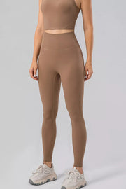 Model wears brown high-waist active leggings with a wide waistband, fitted full-length silhouette, and visible seamed construction.