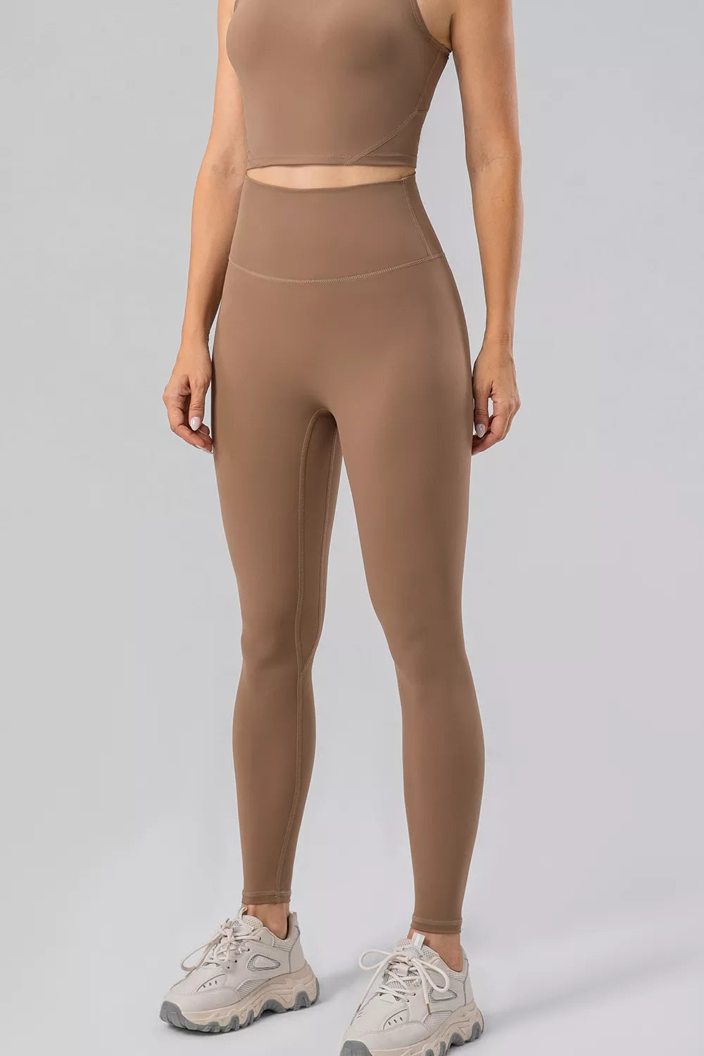 Model wears brown high-waist active leggings with a wide waistband, fitted full-length silhouette, and visible seamed construction.