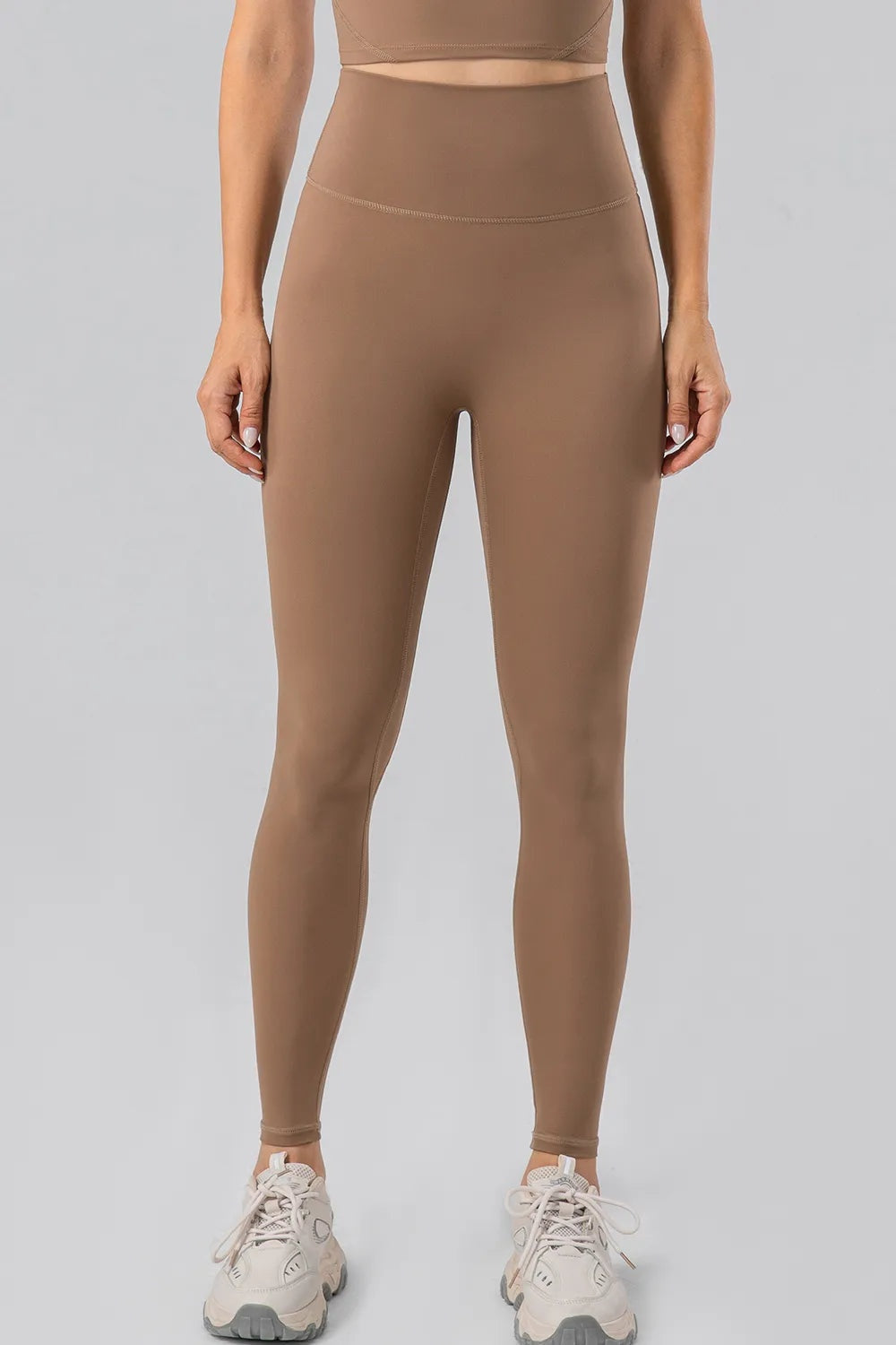 Model stands front-facing in taupe high-waist active leggings with a wide waistband, smooth stretch fabric, fitted silhouette, and ankle-length hems.
