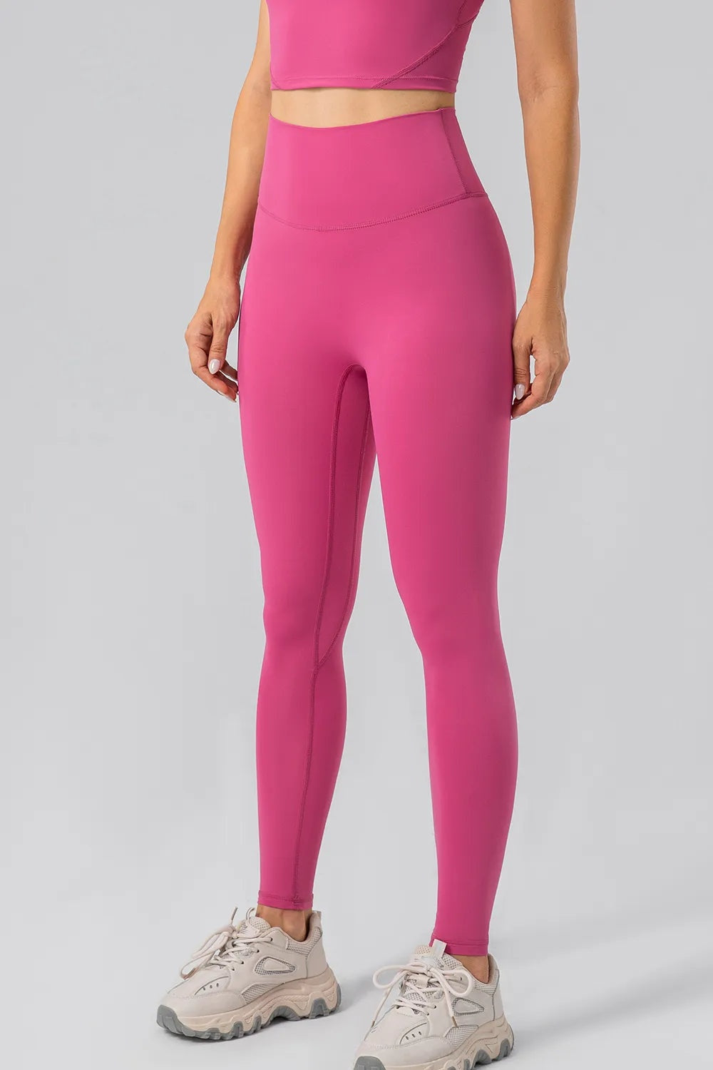 Model standing in bright pink high-waist active leggings with a wide waistband, full-length fitted silhouette, and visible curved seam construction.