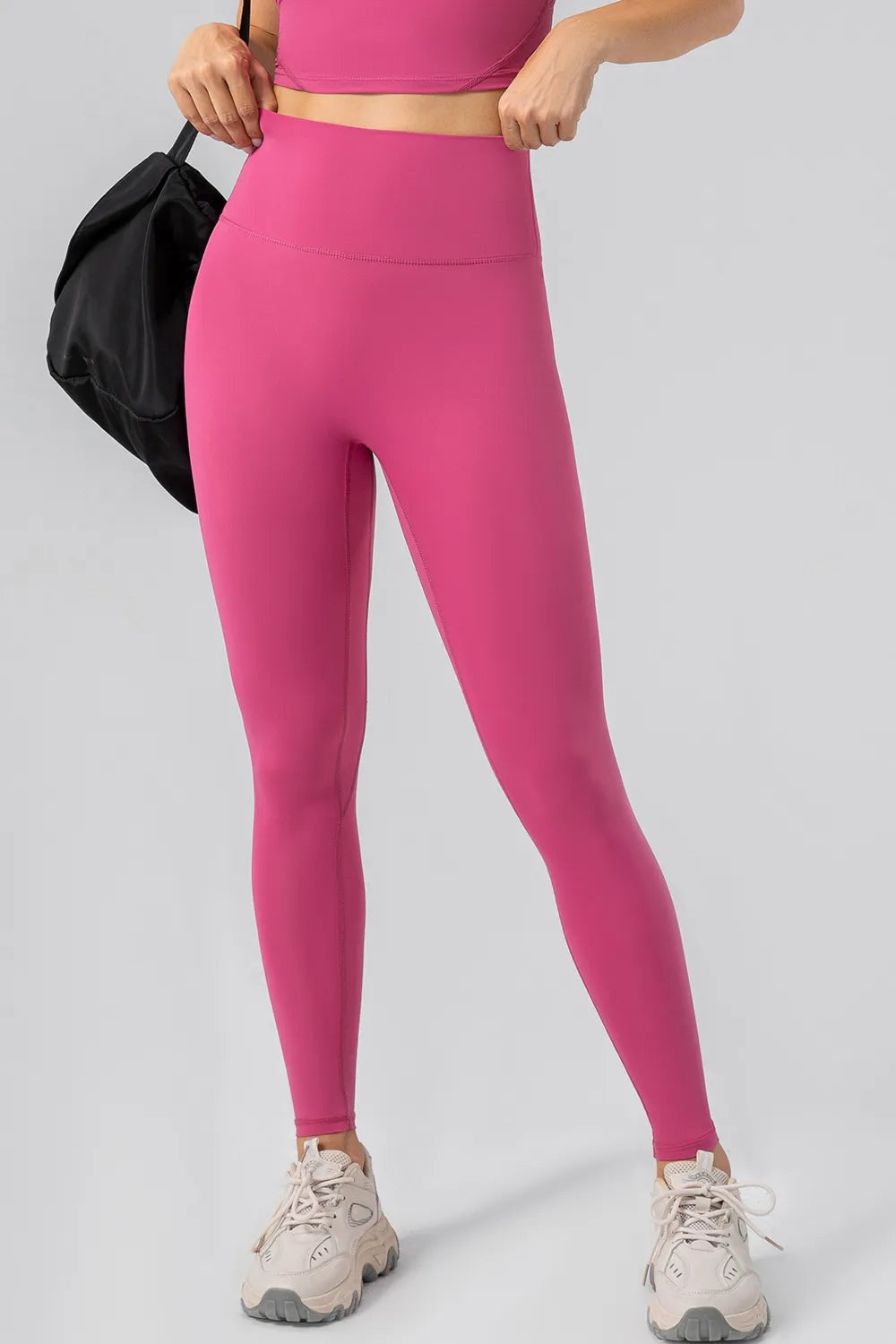 Model adjusts the wide waistband of bright pink high-waist active leggings, showing their sleek, full-length fitted silhouette and visible hip seam.