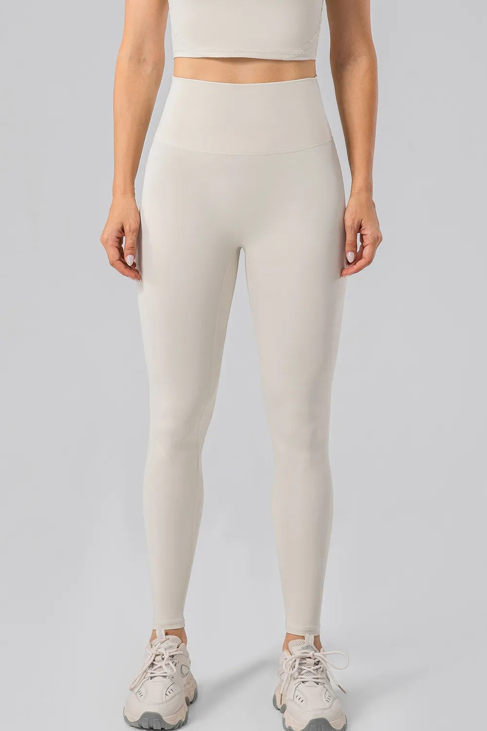 Model stands facing forward in cream high-waist active leggings with a wide waistband, fitted full-length silhouette, and subtle leg seams.