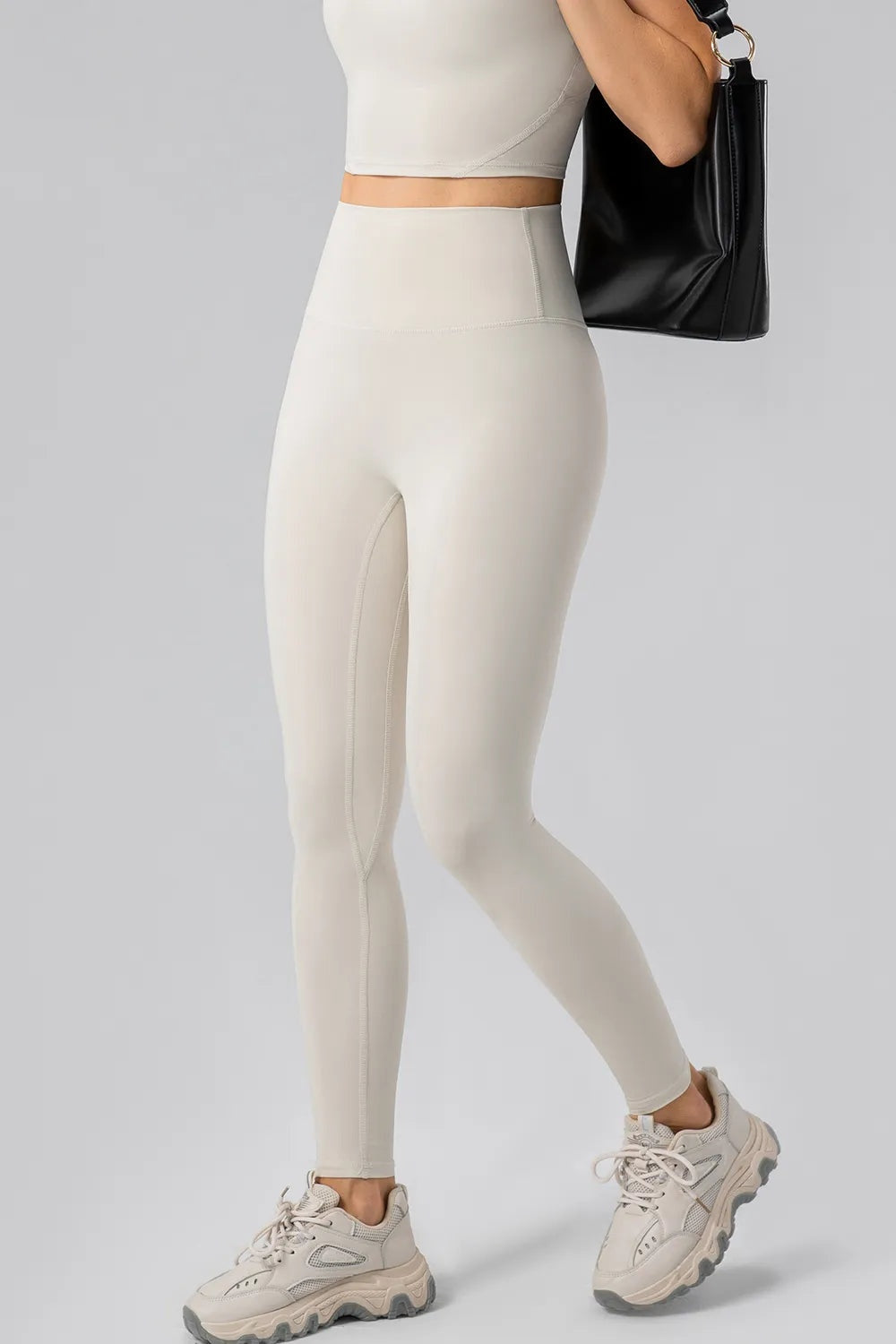 Model wears cream high-waist active leggings with a wide waistband, fitted full-length silhouette, and visible seam construction while posing with one leg extended.