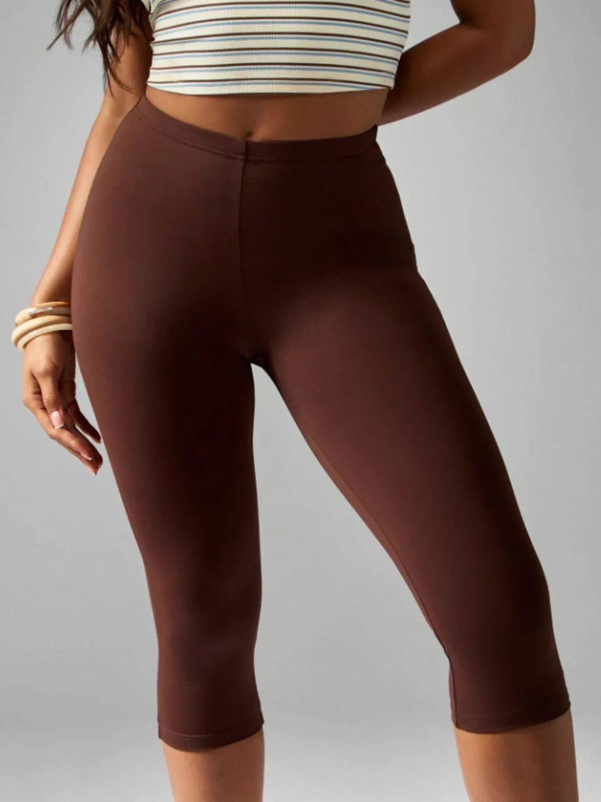 A model wears brown high-waisted active capri leggings with a smooth waistband, fitted stretch silhouette, and clean hems ending below the knees.