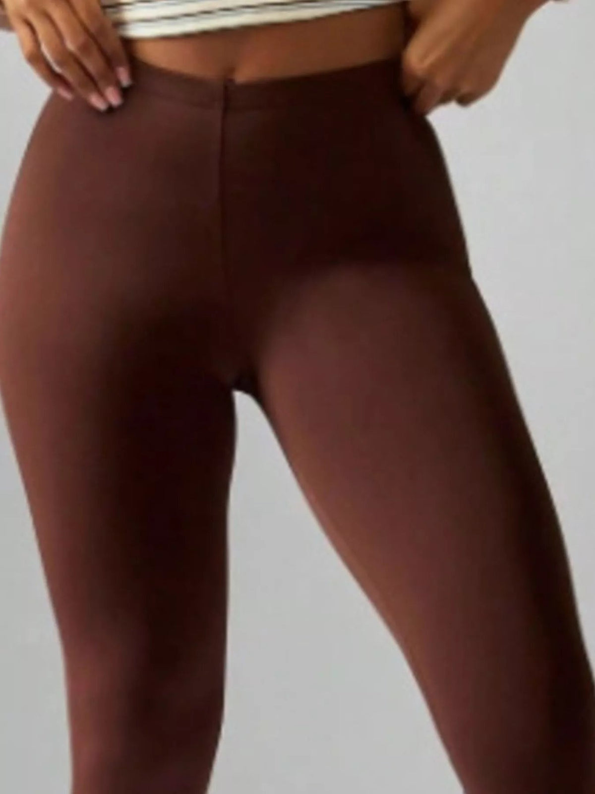 A model adjusts the waistband of dark brown high-waisted active capri leggings, showing their smooth, close-fitting silhouette and center front seam.