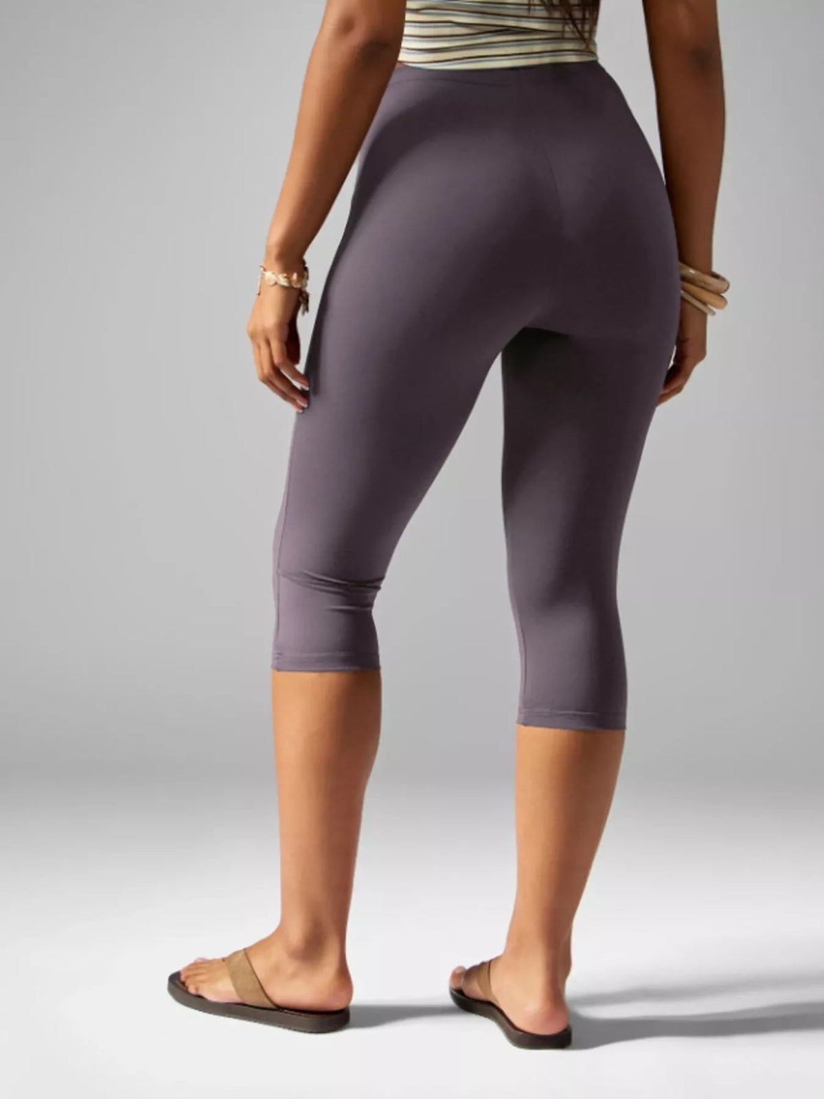 Model shown from behind wearing high-waisted purple active capri leggings with a smooth, close-fitting silhouette and below-knee length.