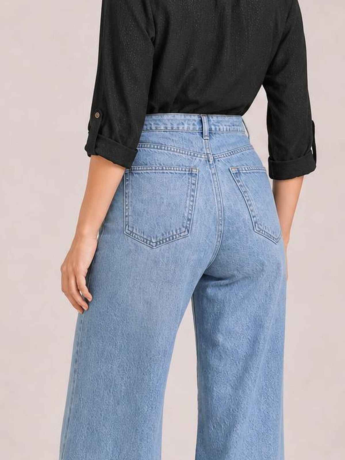 Model viewed from behind wears light blue high-waisted bootcut jeans with belt loops, contrast stitching, and two back patch pockets, posing with one hand on her hip.
