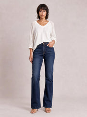 A model stands with one hand in a pocket, wearing dark blue high-waisted bootcut jeans with a button closure, front pockets, and a slim-to-flared silhouette.
