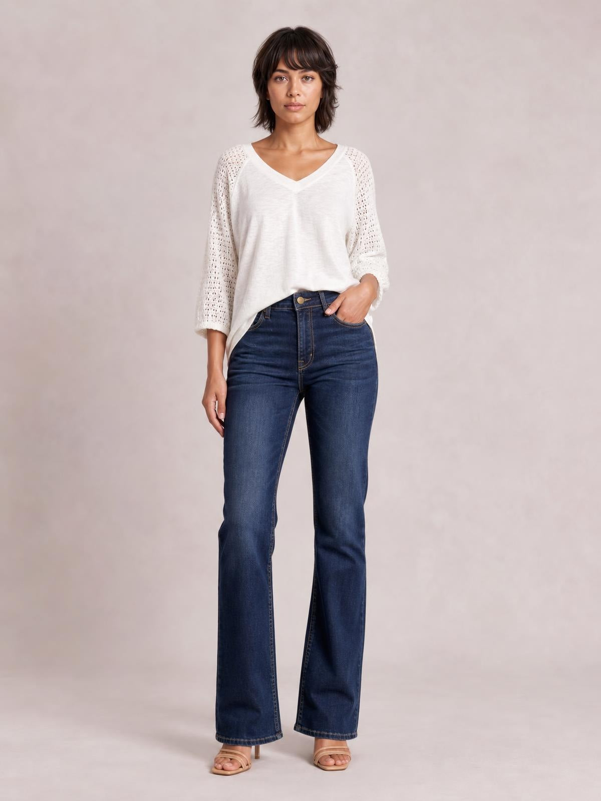 A model stands with one hand in a pocket, wearing dark blue high-waisted bootcut jeans with a button closure, front pockets, and a slim-to-flared silhouette.
