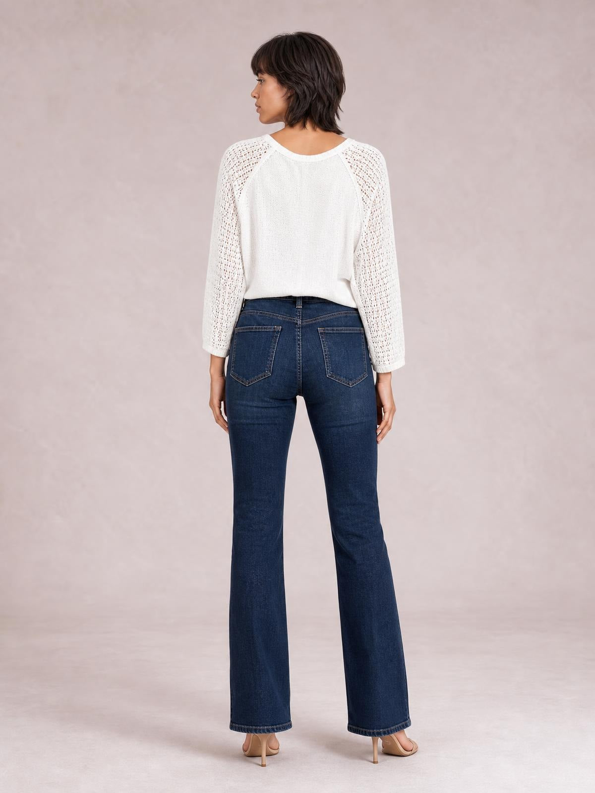 A model stands facing away in dark blue high-waisted bootcut jeans with a fitted silhouette, back pockets, buttoned waistband, and gently flared legs.