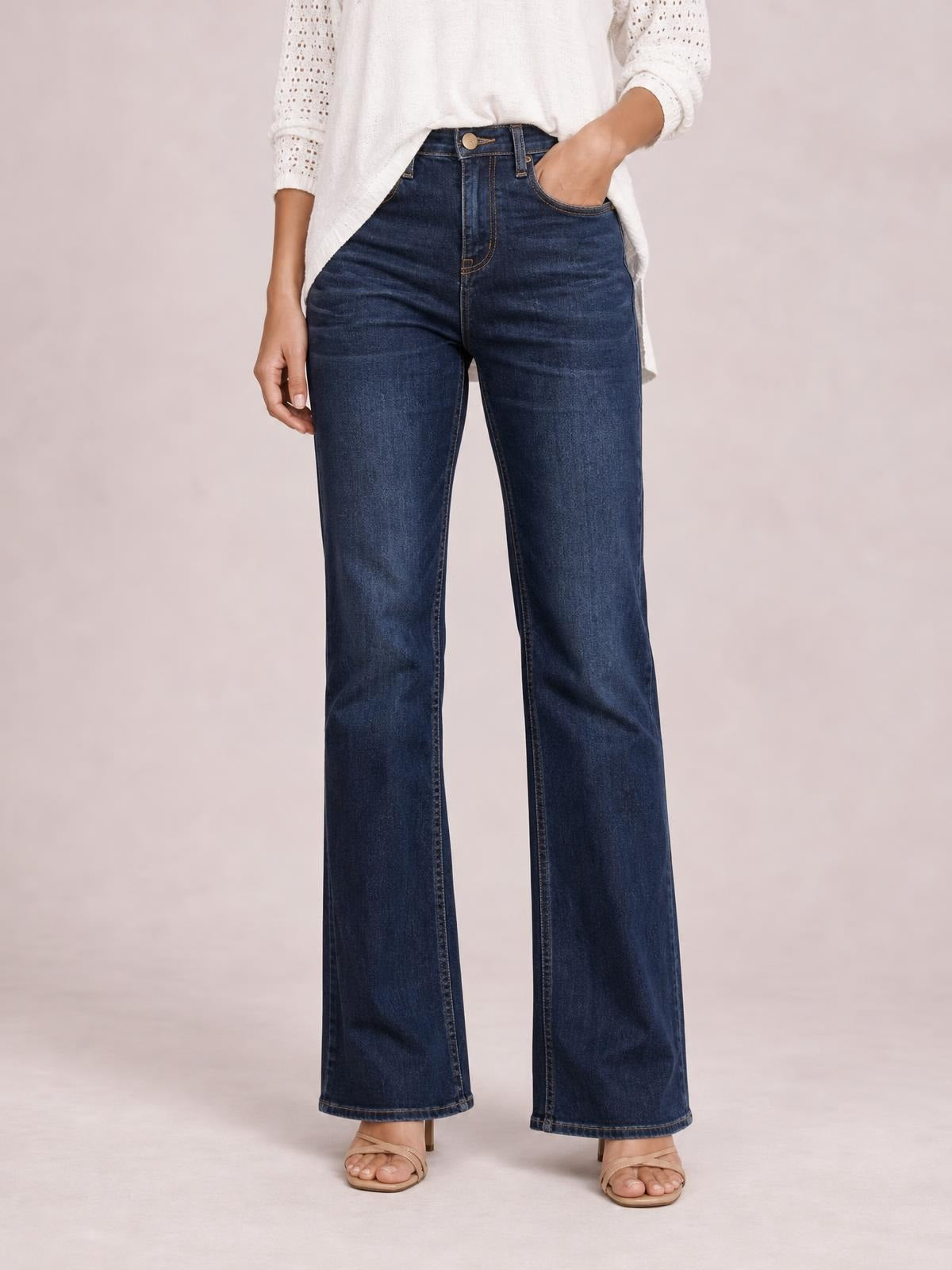 Model stands in dark blue high-waisted bootcut jeans with a button fly, belt loops, front pockets, fitted hips, and subtly flared legs.