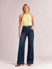 A model stands in dark blue high-waisted flare leg jeans with a button closure, front pockets, wide-leg silhouette, and structured hems, one hand in a pocket.