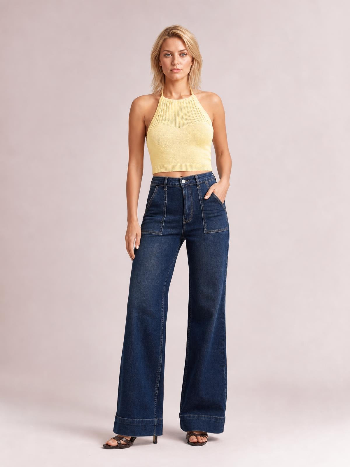 A model stands in dark blue high-waisted flare leg jeans with a button closure, front pockets, wide-leg silhouette, and structured hems, one hand in a pocket.