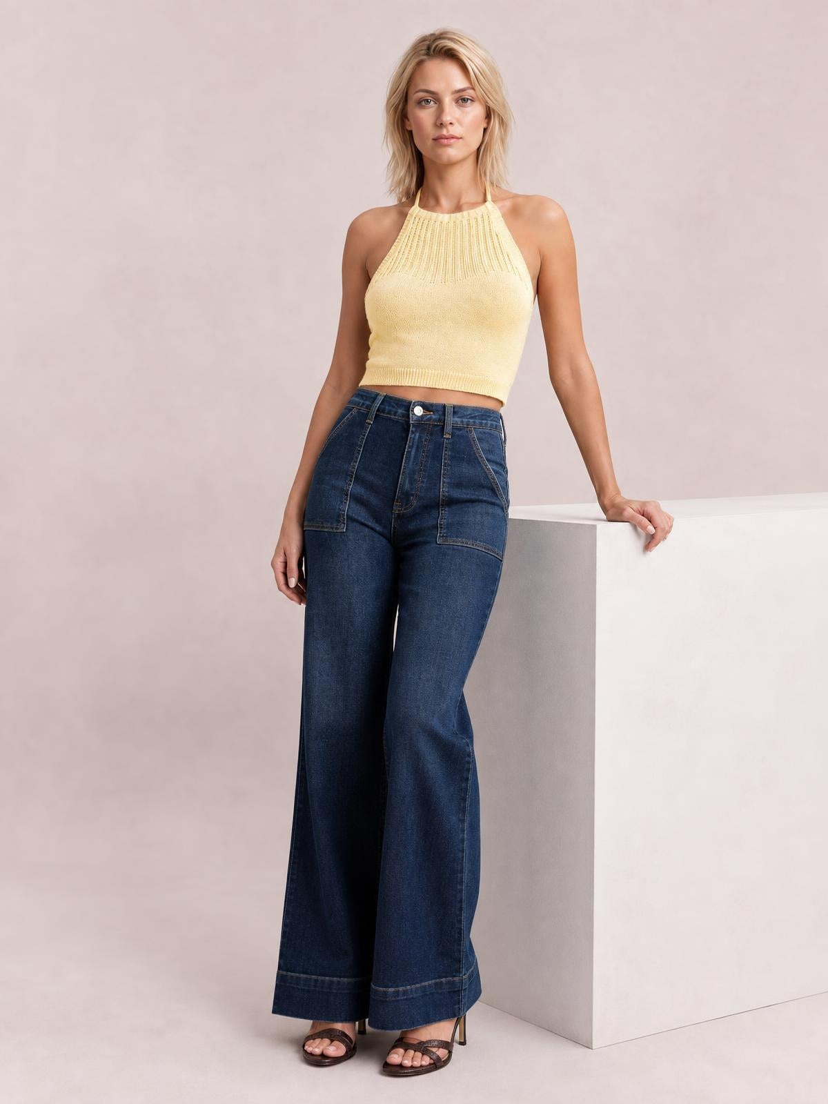 Model stands beside a pedestal wearing dark blue high-waisted flare leg jeans with a button closure, front pockets, and wide full-length legs.