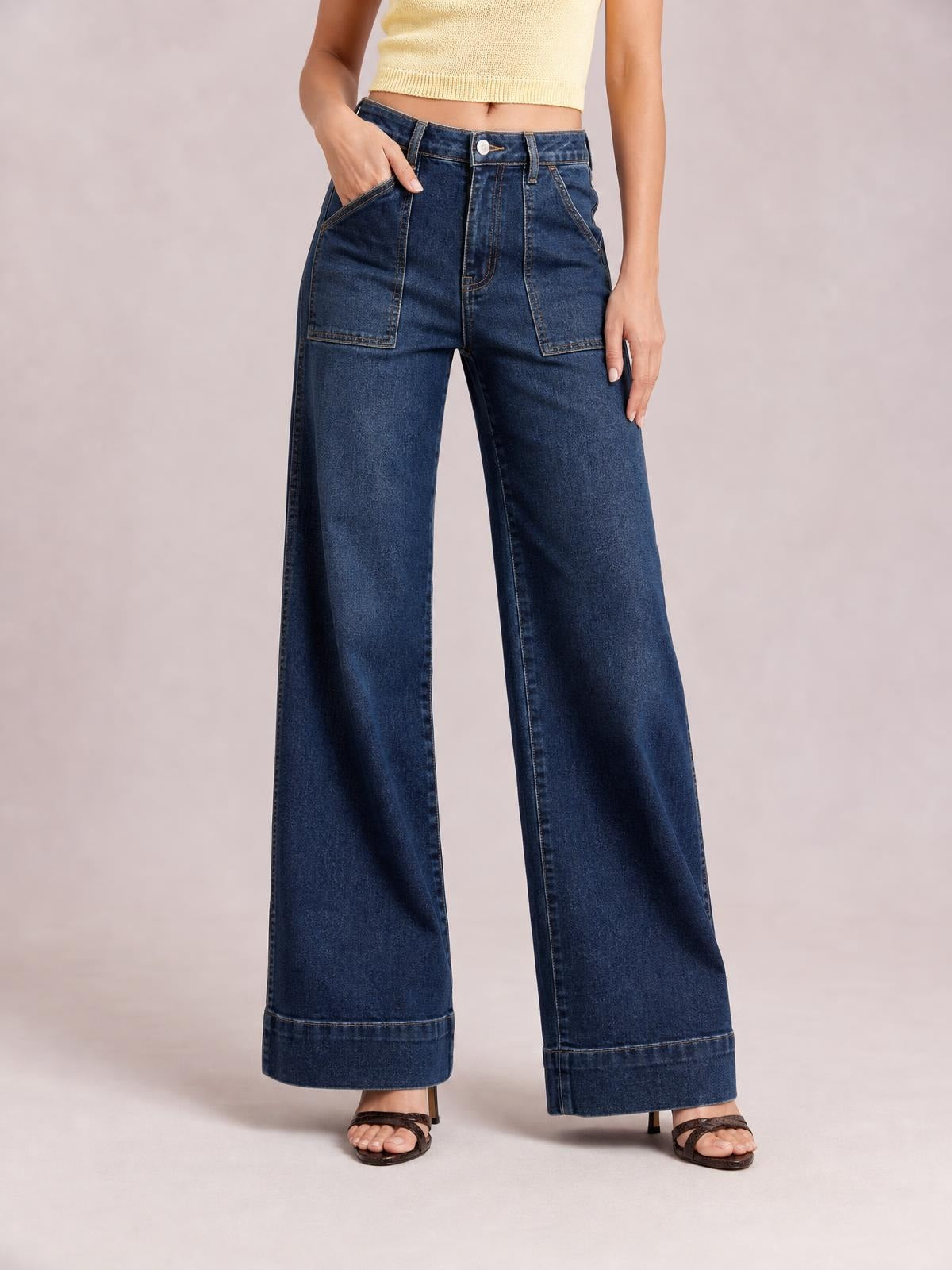A model wears dark blue high-waisted flare leg jeans with a button fly, large front patch pockets, wide legs, and structured hem bands.