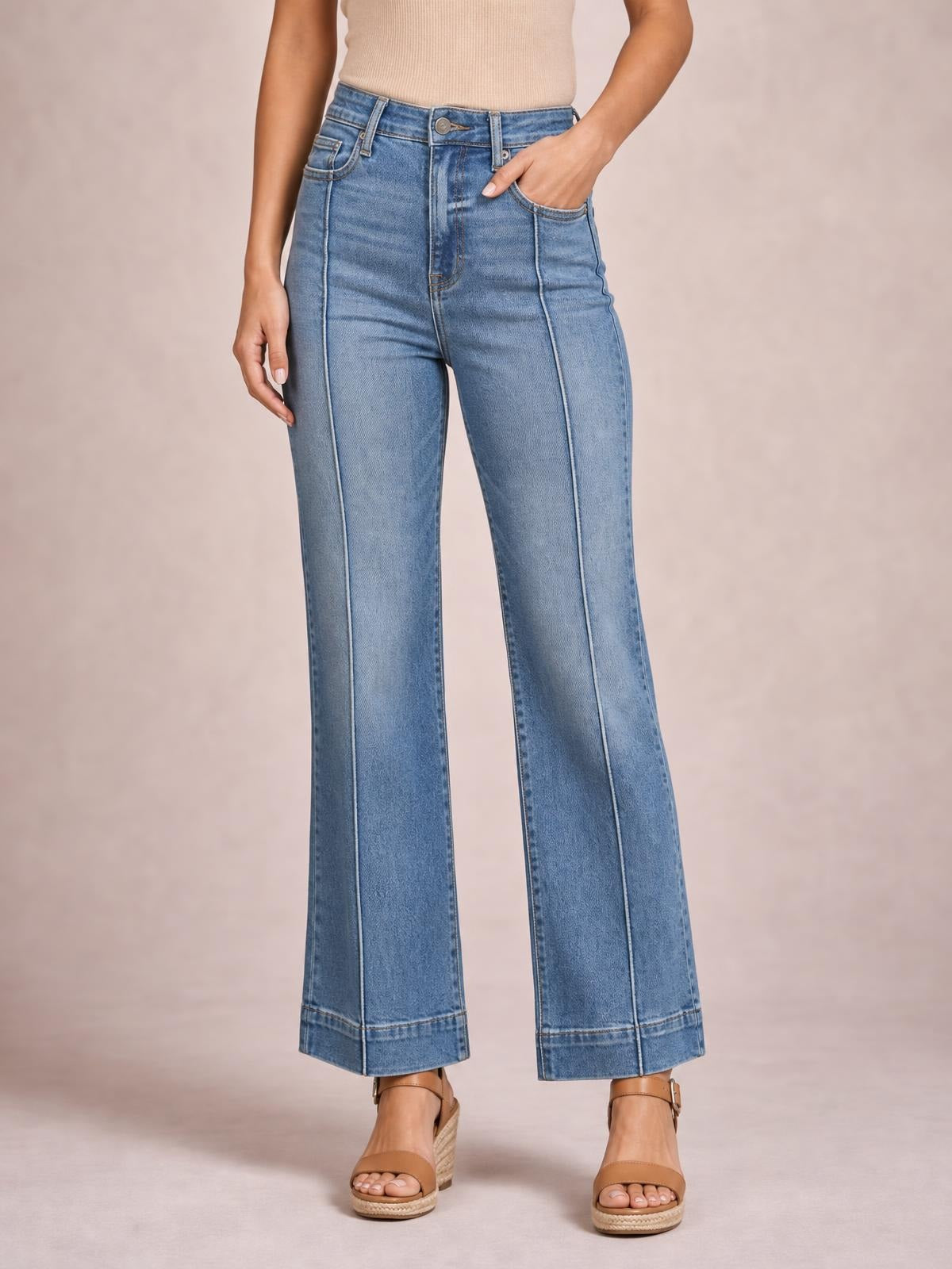 A model wears light blue high-waisted flare leg jeans with a button closure, front and back pockets, vertical seam detailing, and wide hems, posing with one hand in a pocket.