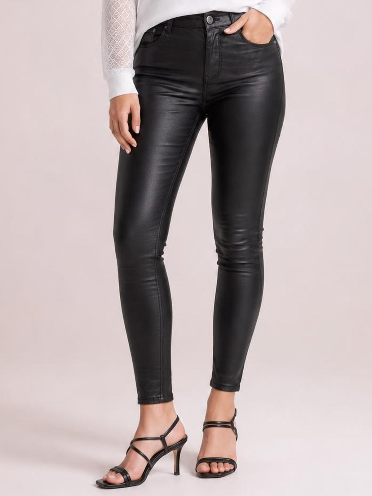 Model wears black high-waisted faux leather skinny pants with front pockets, belt loops, button closure, and a sleek fitted silhouette, posing with one hand in a pocket.