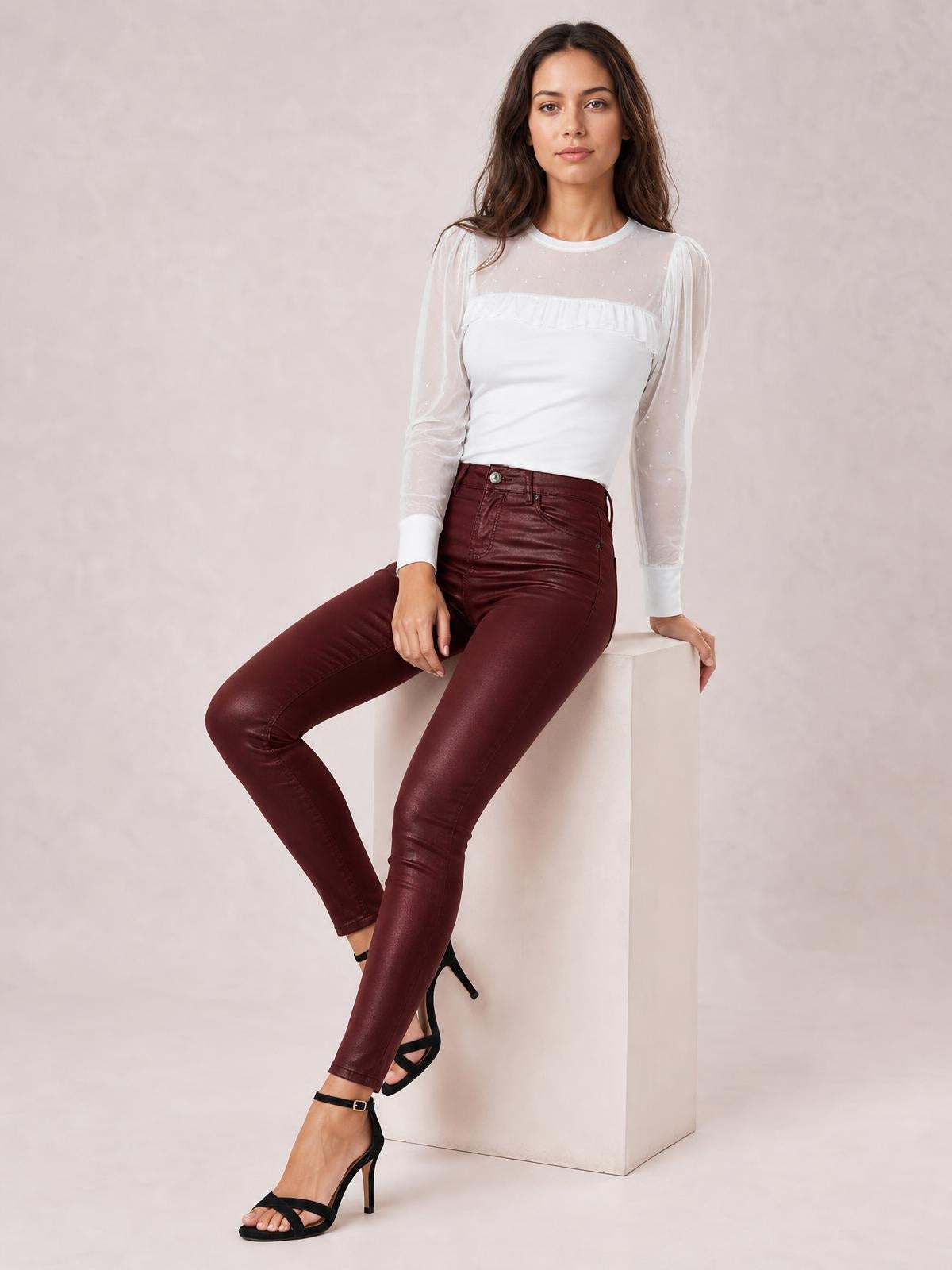 A seated model wears burgundy high-waisted faux leather skinny pants with a sleek fitted silhouette, front pockets, button closure, and visible seam details.