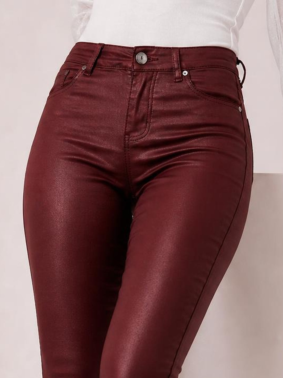 Model wears high-waisted burgundy faux leather skinny pants with a button closure, belt loops, front pockets, and a sleek fitted silhouette.