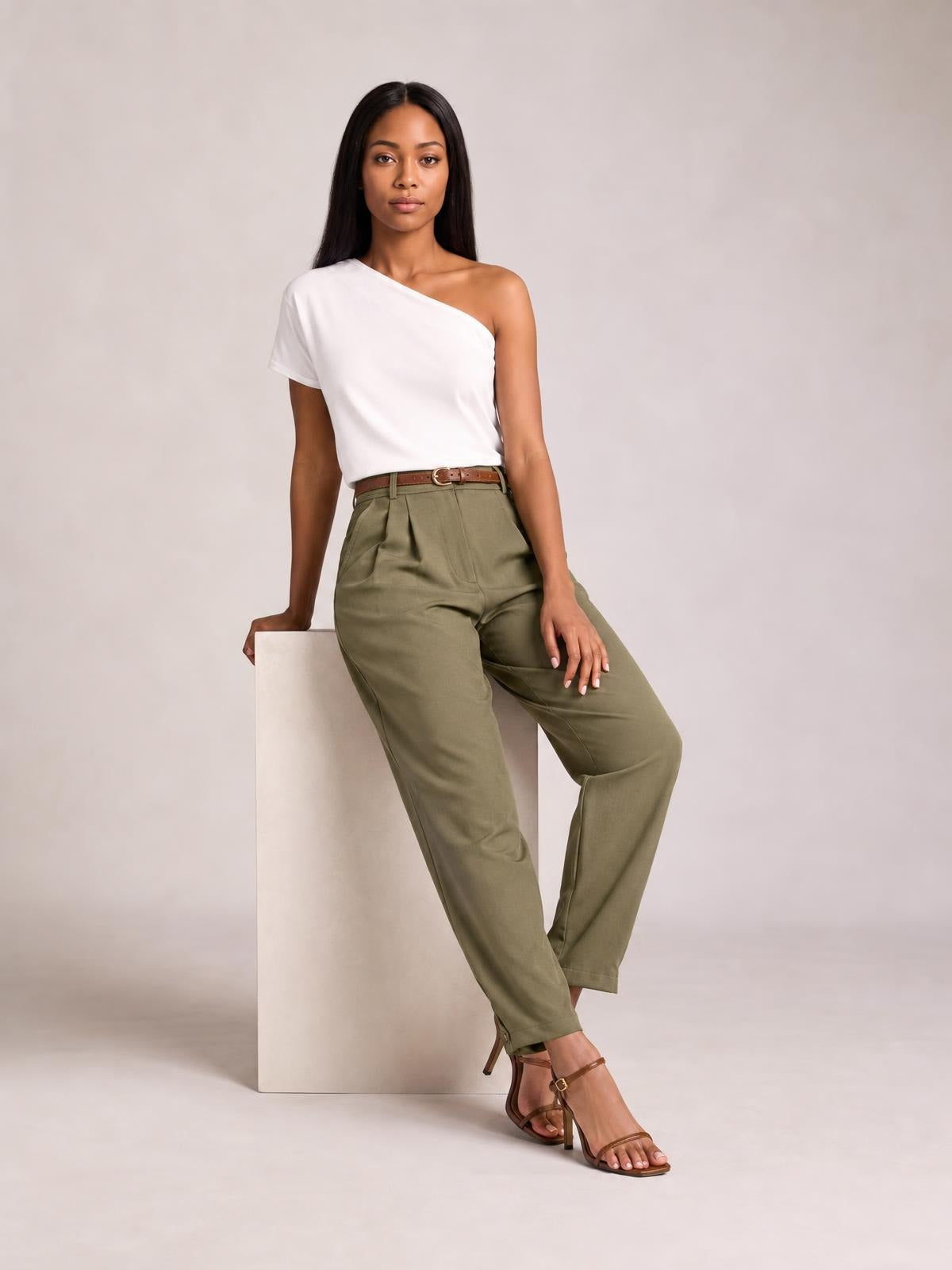 A model wears olive green high-waisted pleated pants with front pleats, side pockets, and a relaxed tapered silhouette while seated with one leg crossed.