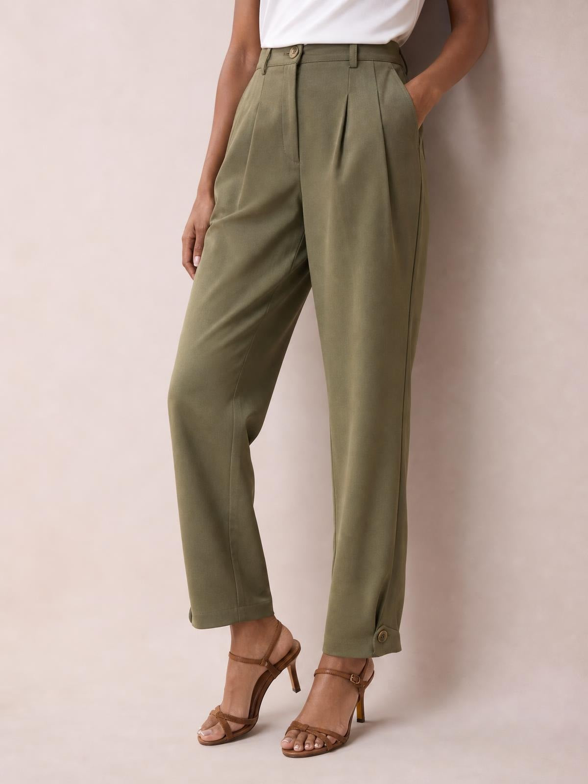 Model wearing olive green high-waisted pleated pants with a relaxed straight-leg silhouette, side pockets, belt loops, and buttoned cuffs, posing with one hand in a pocket.