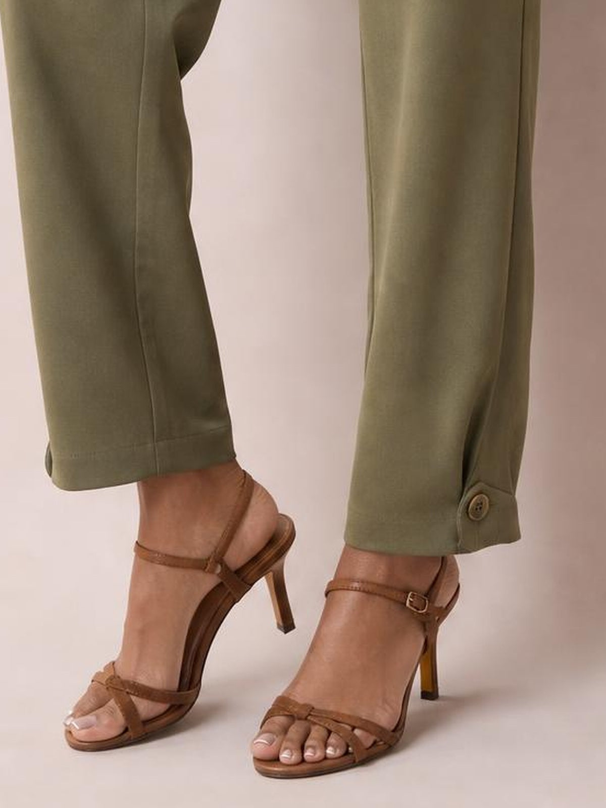A model stands in olive-green ankle-length pants with straight legs, stitched hems, and buttoned ankle tabs, paired with brown strappy heels.