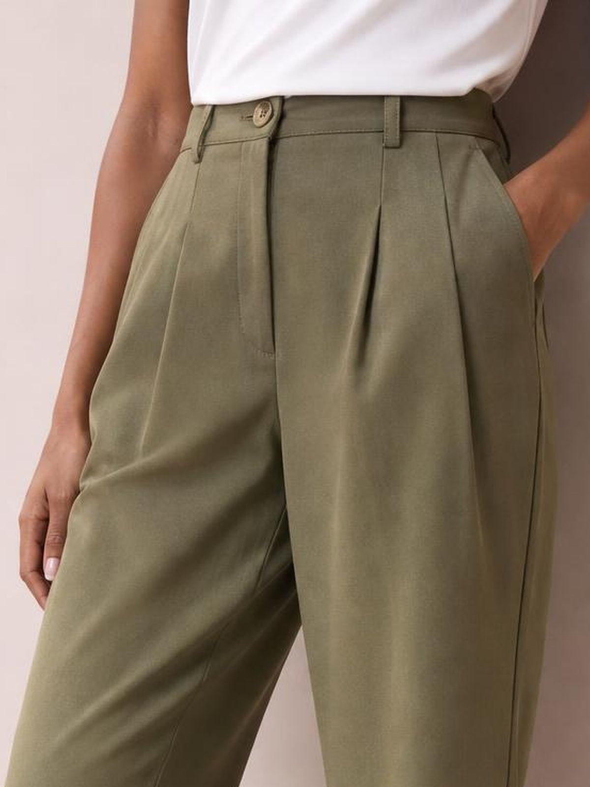 A model wears olive-green high-waisted pleated pants with a relaxed wide-leg silhouette, belt loops, button closure, and side pockets, with one hand tucked in.