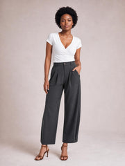 Model stands in dark gray high-waisted pleated pants with a wide-leg silhouette, front button closure, and side pockets, posing with one hand in a pocket.