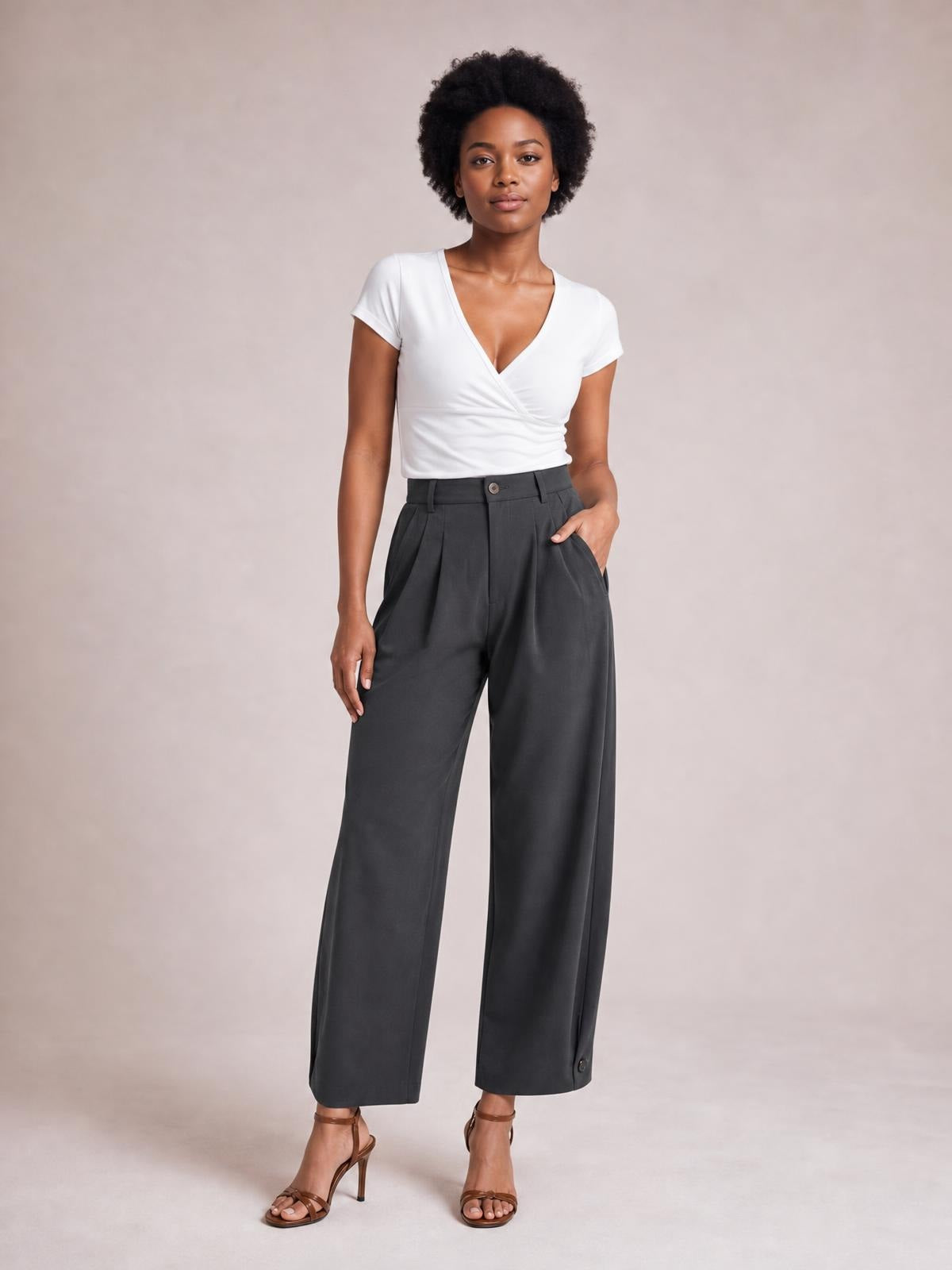 Model stands in dark gray high-waisted pleated pants with a wide-leg silhouette, front button closure, and side pockets, posing with one hand in a pocket.